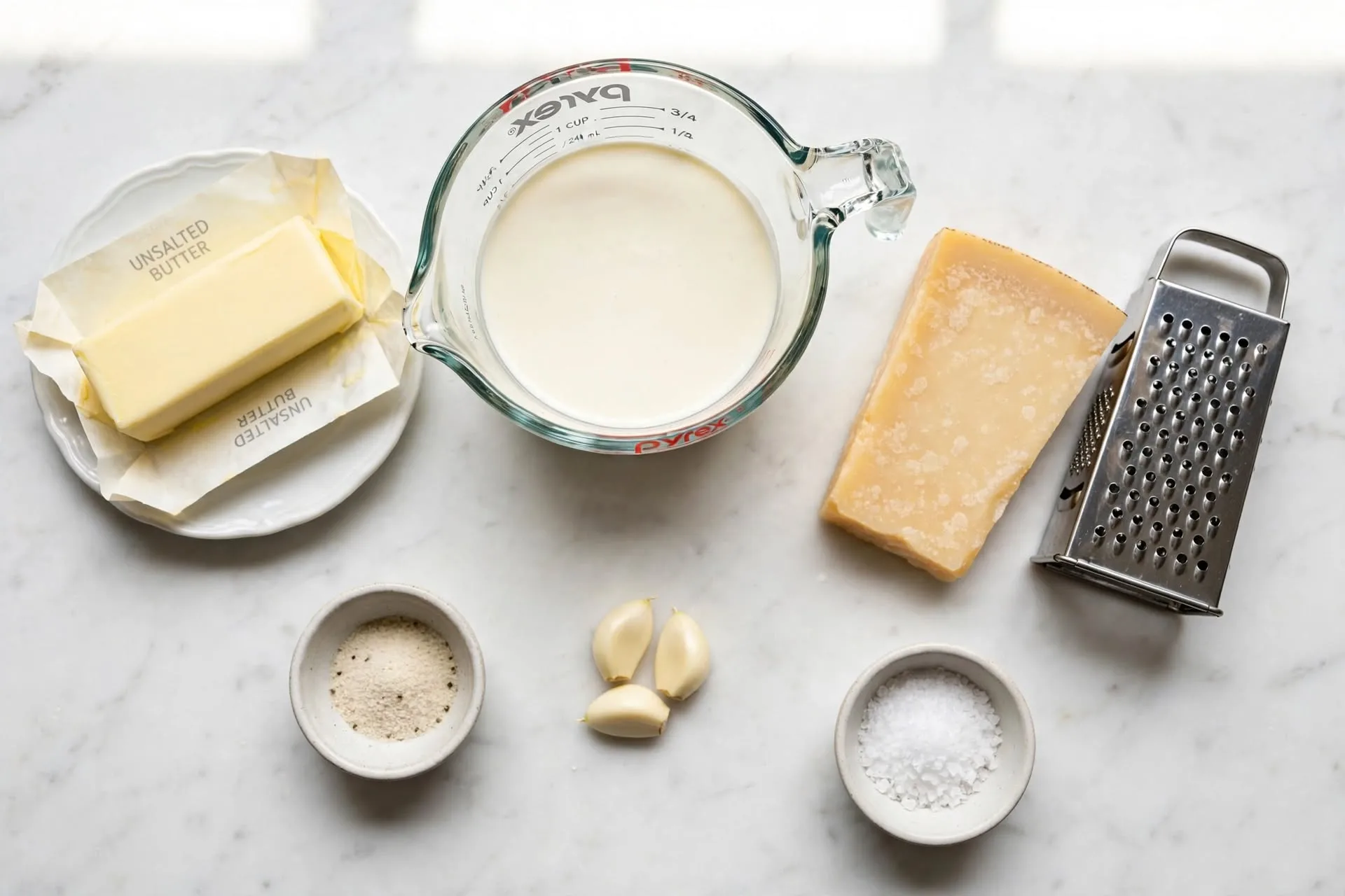 All ingredients for homemade alfredo sauce laid out on white marble including butter, heavy cream, Parmesan, garlic, white pepper, and salt, homemade alfredo sauce ingredients